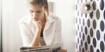 Une femme réfléchit assise aux toilettes à côté d’un mur à pois et d’un rouleau de papier toilette. 