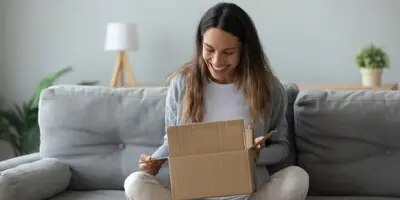 Une femme est sur un canapé gris, elle arbore un grand sourire en regardant à l’intérieur de son colis surprise.